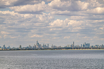 Fototapeta premium New York, NY, USA - August 1, 2023: City skyline off ocean under blue cloudscape. One World Trade center is tallest. Green roof is Federal Hall. Bel Harbor sandy beach line