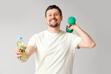 Sporty young man with bottle of cucumber water and dumbbell on light background