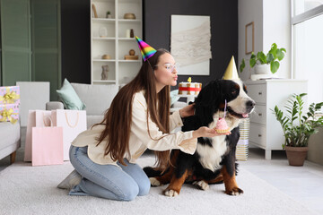 Young woman and cute Bernese mountain dog with party hats and cake at home. Birthday celebration