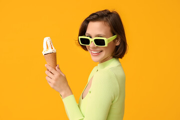 Beautiful young woman in sunglasses with sweet ice-cream in waffle cone on yellow background