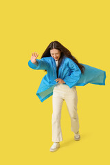 Young Asian woman in raincoat with strong wind on yellow background