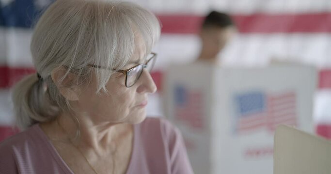 Senior woman wearing spectacles and hair up, considers options while casting votes at booth with US flag on wall behind. Gimbal move to from medium shot to BCU. Asian man votes in background