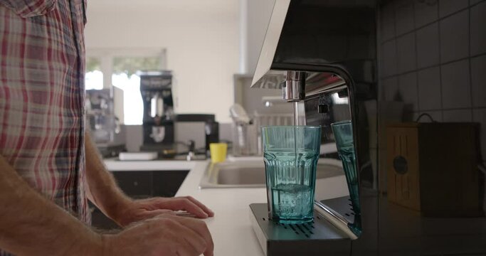 A man is pouring water from a modern filtration device into a clear glass, emphasizing a healthy lifestyle choice in his well-lit kitchen.