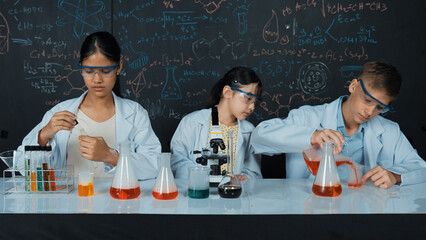 Cute girl looking under microscope while student doing experiment at blackboard with theory...