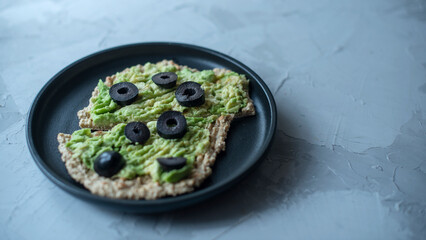 avocado toast on slices of toasted bread topped with ground lemon pepper and olives on a grey stone background, copy space