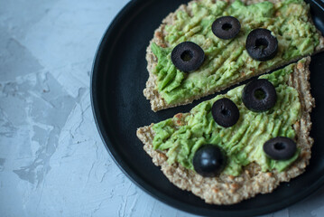 avocado toast on slices of toasted bread topped with ground lemon pepper and olives on a grey stone...