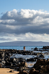 Two women in bikinis standing on black lava rocks jutting out into the Pacific Ocean at low tide,...