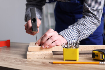Professional repairman hammering nail into board at wooden table indoors, closeup