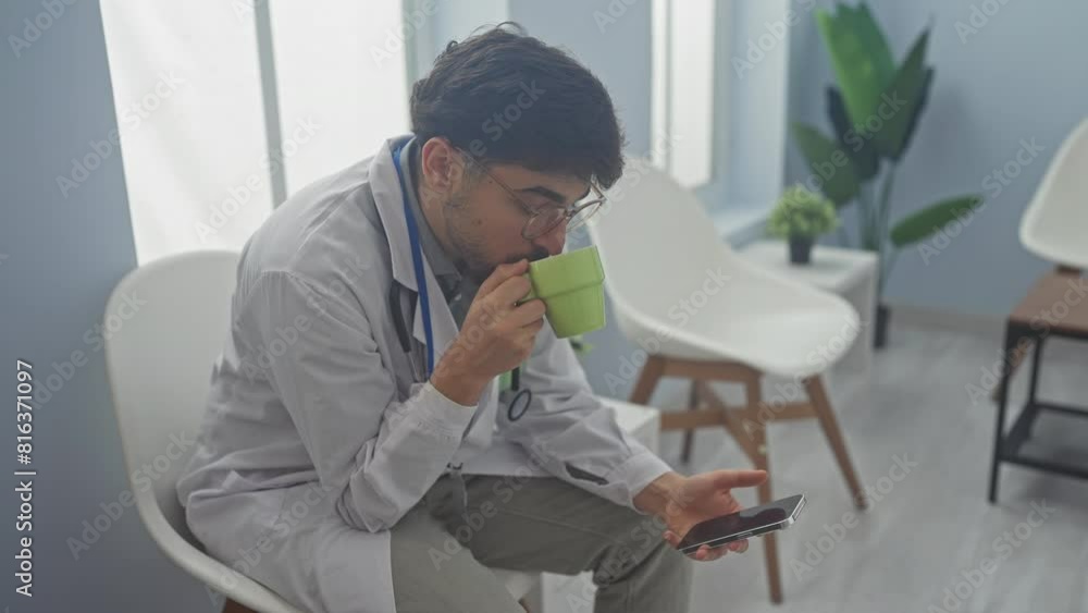 Poster a young bearded male doctor in glasses, using a smartphone and drinking coffee while taking a break 