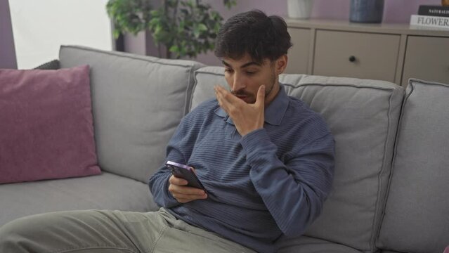 Shocked young arab man covering mouth with hand, a saucer-eyed surprise sitting on sofa at home, afraid and speechless after viewing shocking news on smartphone