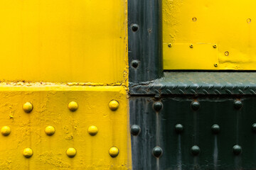 Fading black and yellow paint on heavy metal wall. Large aluminum or steel rivets fasten the material together. This train car structure sides outside and corrodes over time