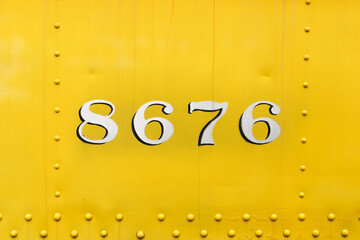 8676, stencil painted numbers on a vintage, antique train car. Yellow paint on thick sheet metal and four digits on a very old locomotive. Rivet rows surround the symbol