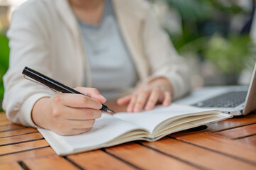 A cropped image of a woman working remotely at a cafe outdoor table, taking notes in her book.