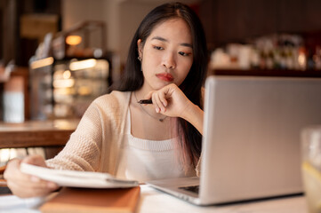 Young focused Asian woman working remotely at a coffee shop, taking notes in her notebook.