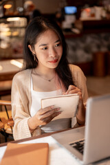 Fototapeta premium A focused female college student sits in a coffee shop taking notes, studying online on her laptop.