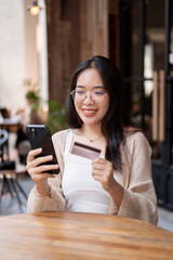 A happy Asian woman sits at an outdoor table of a cafe, holding her credit card and smartphone.