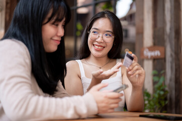 Two happy Asian female friends are talking about the benefits of their credit cards at a cafe.