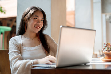 A beautiful, happy Asian woman is sitting outdoors, working remotely at a cafe, using her laptop.