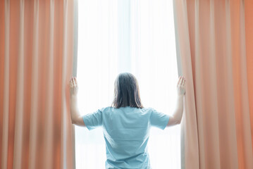 Young woman stands at window and opens curtains, back view.