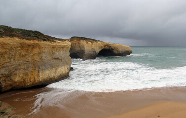 London Arch rock formation with ocean waves, sandy beach and cloudy sky on the Great Ocean Road in Victoria, Australia