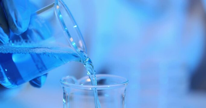 Blue liquid is poured from a glass beaker into a flask in laboratory