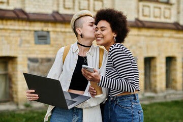 Two young women in casual attire stand in front of a building, working on a laptop.