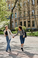 Obraz premium Two young women, holding hands, walk down a cobblestone street near a university campus.