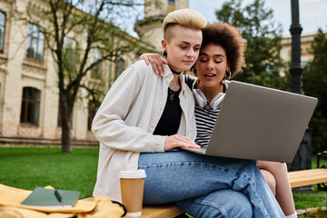 Two young women in casual clothing sitting on a bench, engrossed in their laptop screen.