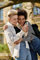 Two young women in casual attire embrace each other while engrossed in their cell phones, seemingly...