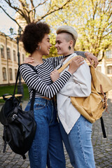 Obraz premium Two young women, one Black and one Caucasian, embrace each other warmly while holding backpacks outdoors.