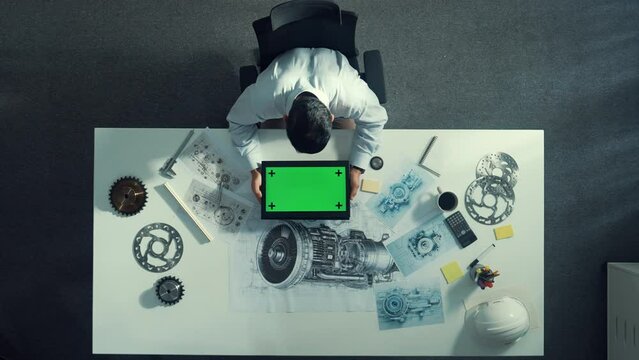 Top view of skilled engineer writing in paper while looking at tablet display green screen with turbine engine sketch design and metal gear and architectural equipment scatter around. Alimentation.