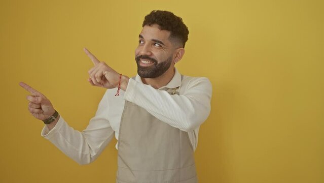 Young hispanic man wearing apron standing smiling and looking at the camera pointing with two hands and fingers to the side. over isolated yellow background