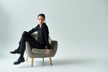 A young queer person gracefully poses on a chair in a studio against a grey backdrop.