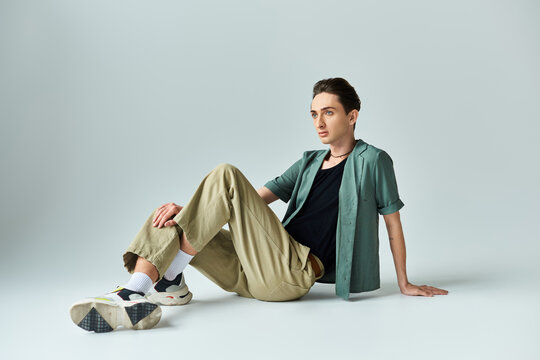 A young queer person in a tan shirt and khaki pants sits on the floor in a thoughtful pose in a studio setting against a grey background. - Powered by Adobe