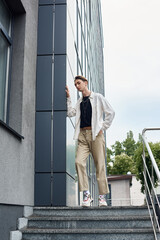 A young queer person in stylish attire stands confidently on the steps of a building celebrating LGBT pride.