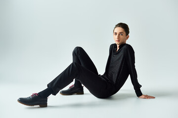 A young queer person is seated on the floor, wearing a black sweater, striking a pose in a studio...