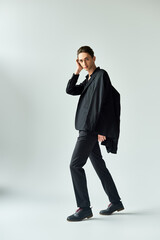 A young queer person strikes a pose in a studio, donning a stylish black jacket and pants, against a grey background.