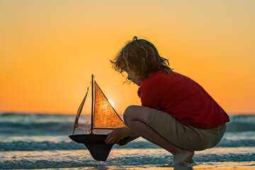Kid playing with toy sailboat on sea beach at the summer sunset time. Sunset silhouette of child...
