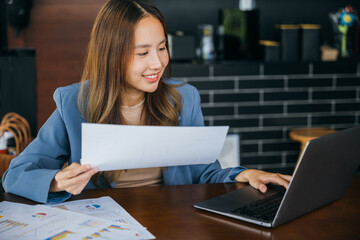 Fototapeta premium Happy accounting female reviewing data in financial charts and graphs outside at cafe coffee shop, smiling young business woman reading report paperwork document to analyse with laptop computer