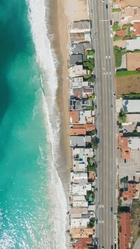 Vertical Screen: Dramatic Aerial Shot of Turquoise Waves Crashing on the Sandy Beach of Malibu, California, Framed by Coastal Homes. 4K Footage. 