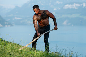 Muscular man using scythe. Farmer with a scythe on green grass field. Farmer cut grass. Rural...