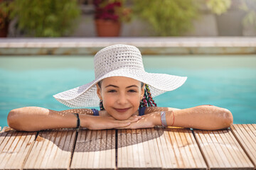 Little smiling girl with white straw hat is leaning on the edge of the pool looking at the camera.  Horizontally. 