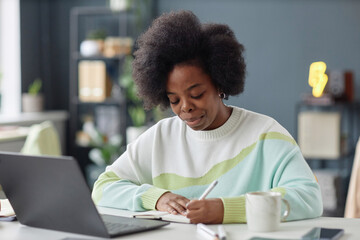 Portrait of Black woman with natural curly hair working at desk in office and writing notes in...