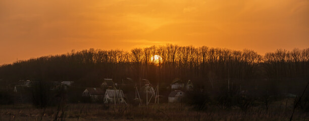Panoramic view of the landscape with the setting Sun behind the trees on the horizon. Yellow-orange sunset over a forest and meadow in the countryside