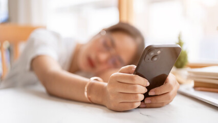 A tired, bored Asian woman leaning on the table and holding her smartphone, waiting for the messages