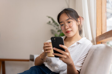 A woman reading online blog on her smartphone while sipping coffee on the sofa in the living room.
