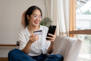 A young, cheerful Asian woman sits on a sofa, using a mobile banking app, shopping online at home.