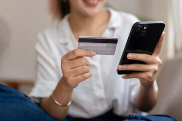 A young, cheerful Asian woman sits on a sofa, using a mobile banking app, shopping online at home.