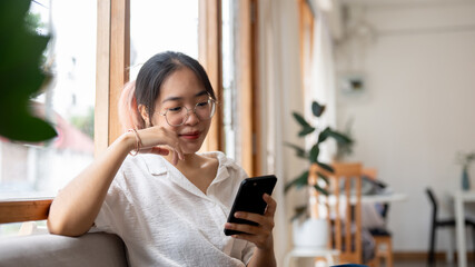 Obraz premium A young, positive Asian woman using her smartphone on a sofa in the minimalist cafe.