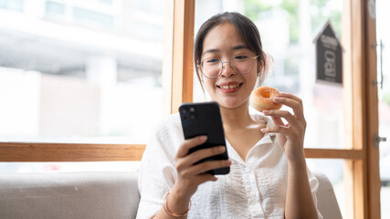 Fototapeta premium A young, happy Asian woman enjoying a donut while using her smartphone on a sofa in a cafe.